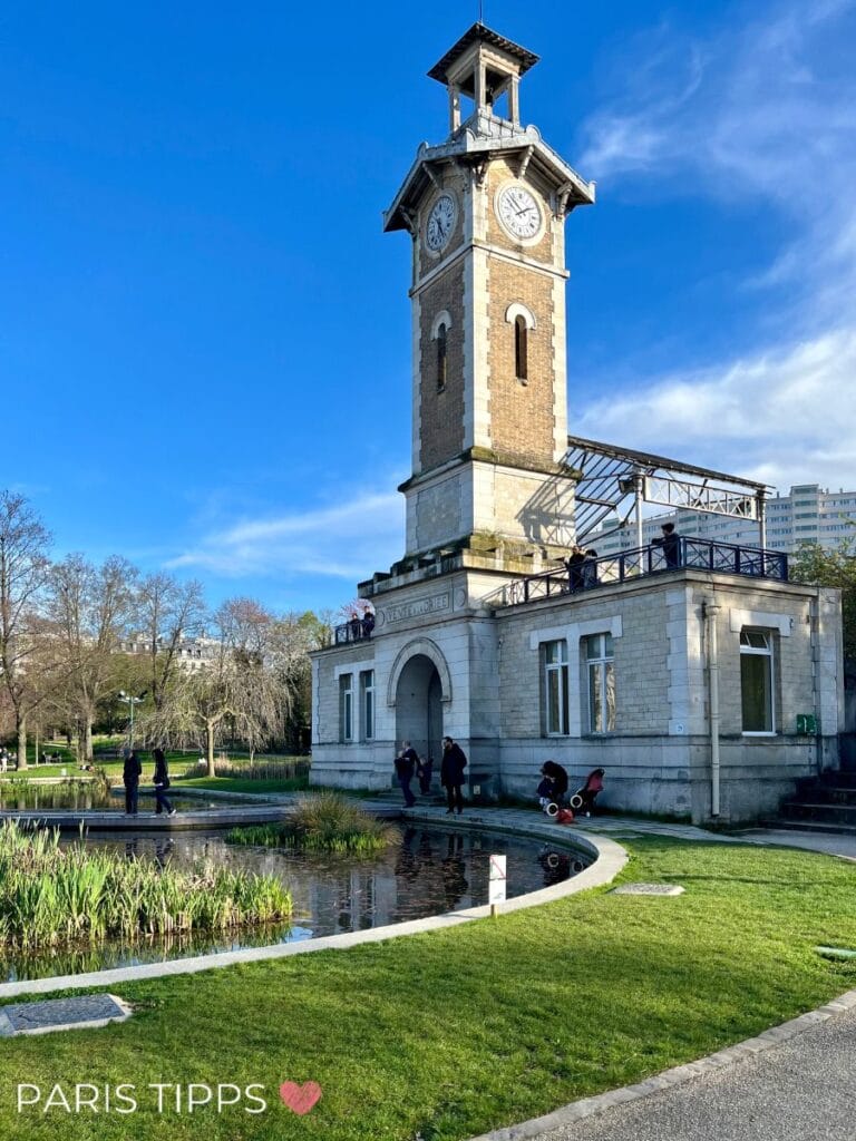 Der Uhrturm im Parc Georges Brassens in Paris, umgeben von gepflegtem Grün, einem kleinen Teich und Spaziergängern, unter strahlend blauem Himmel.