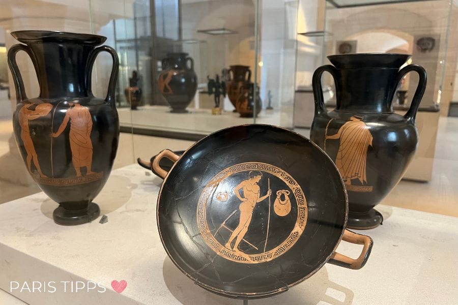 Antike griechische Keramik im Petit Palais in Paris, darunter eine Schale und zwei Vasen mit rotfigurigen Darstellungen von Menschen und mythologischen Szenen. Die detailreichen Verzierungen zeigen klassische griechische Kunstfertigkeit.