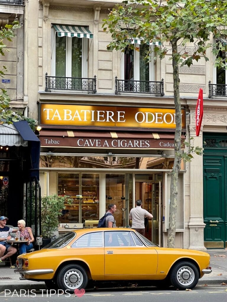 Gelbes Oldtimer-Auto vor der Tabatière Odéon, einem charmanten Tabakladen mit Zigarren-Cave, im Pariser Stil; daneben ein Café mit Menschen, die das Straßenleben genießen.