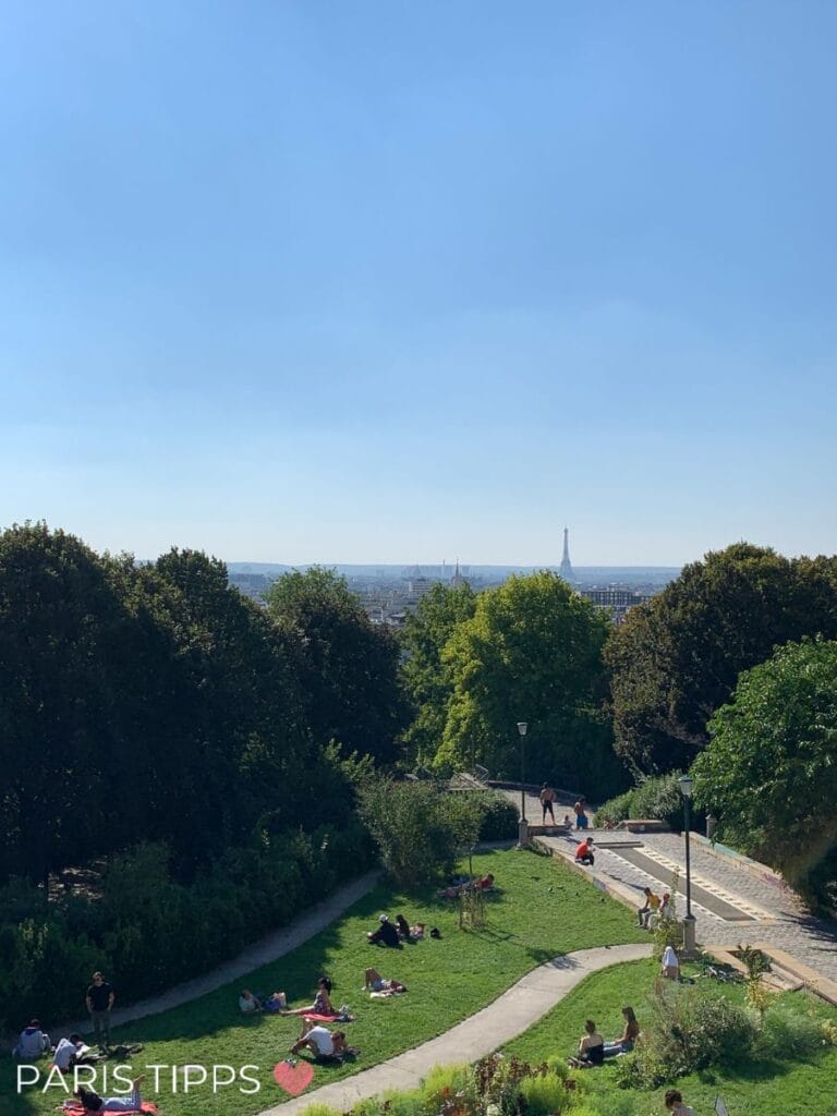 Grüne Wiese im Parc de Belleville in Paris mit entspannten Menschen beim Picknicken und einem Blick auf die Skyline der Stadt, inklusive Eiffelturm in der Ferne.