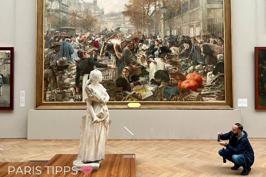 Kunstgalerie im Petit Palais in Paris mit dem beeindruckenden Gemälde „Les Halles“ von Léon Augustin Lhermitte, das einen lebhaften Marktplatz zeigt. Im Vordergrund steht eine weiße Marmorskulptur, während ein Besucher das Kunstwerk fotografiert.