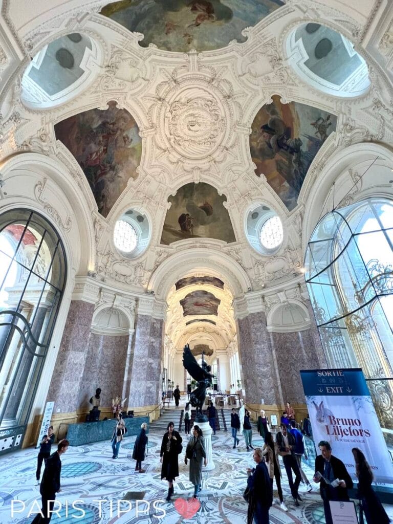 Innenansicht des Petit Palais in Paris mit kunstvoll verzierten Deckenfresken, Marmorsäulen und einem detailreichen Mosaikboden. Besucher schlendern durch die lichtdurchflutete Eingangshalle, die Eleganz und Kunstgeschichte vereint. Im Hintergrund ist eine Statue zu sehen.