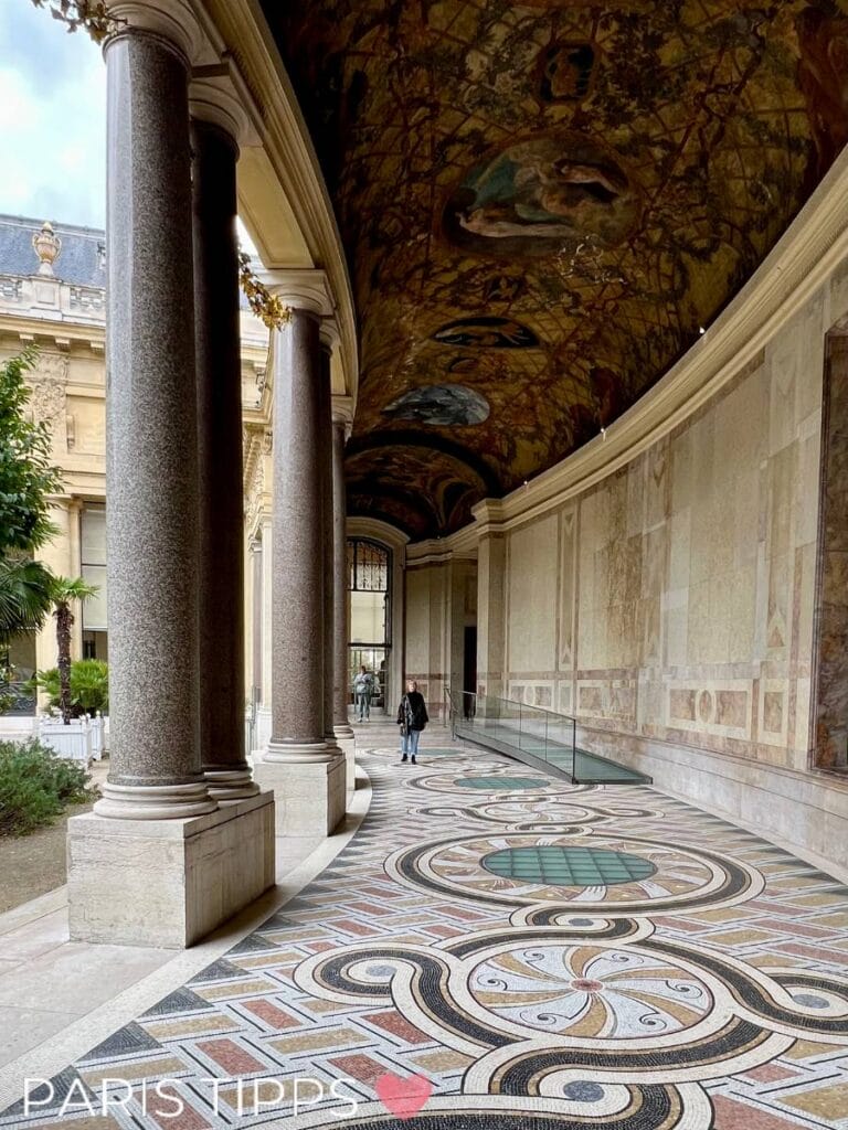 Arkadengang im Petit Palais in Paris mit kunstvoll verzierten Mosaikböden und reich bemalter Decke. Die Säulen aus poliertem Stein rahmen den Gang ein, während im Hintergrund der Blick in den Innenhof führt. Ein Besucher spaziert durch die elegante Passage.
