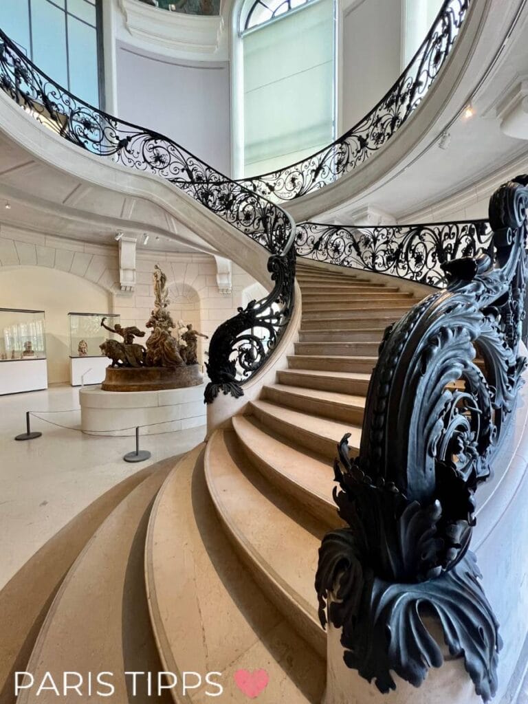 Elegante Wendeltreppe im Petit Palais in Paris mit kunstvoll verziertem schmiedeeisernem Geländer. Die Treppe führt zu einer lichtdurchfluteten oberen Etage, umgeben von großzügigen Fenstern und Kunstwerken im Hintergrund.