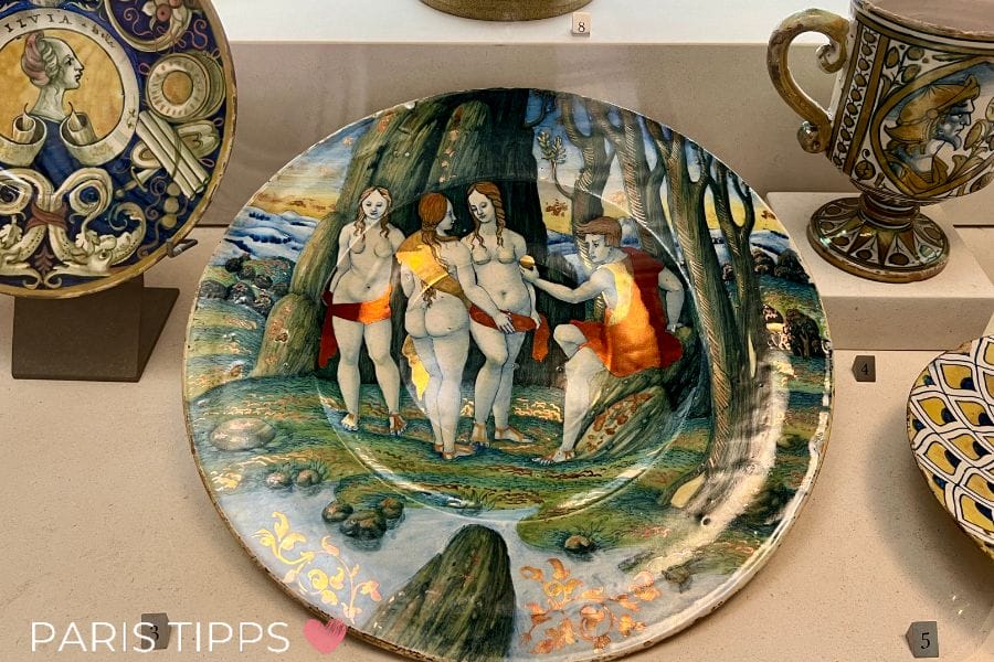 Renaissance-Teller ausgestellt im Petit Palais in Paris, kunstvoll bemalt mit der Szene des Urteils des Paris, in der ein Mann drei Frauen einen goldenen Apfel überreicht. Umgeben von weiteren dekorativen Keramiken.