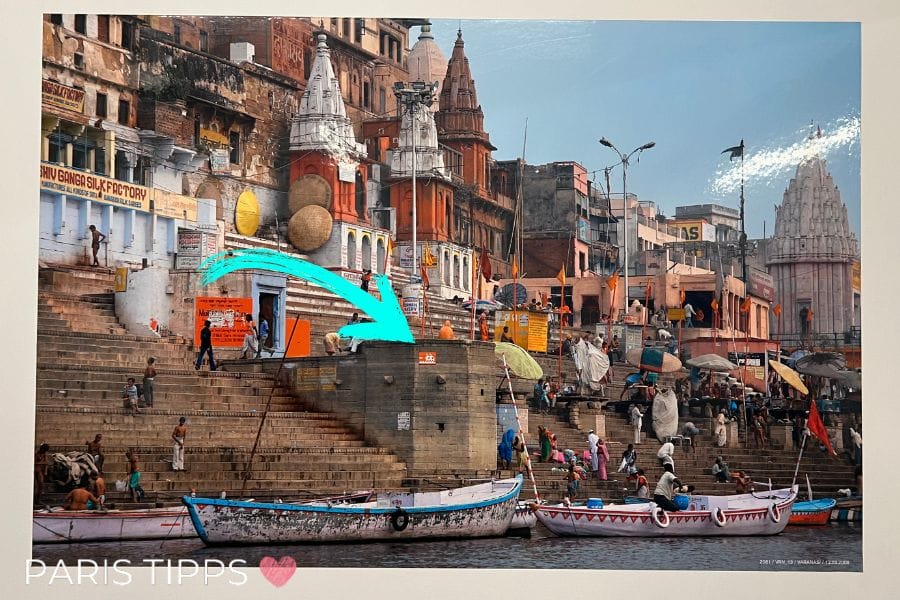 Szene in Indien an einem Fluss. Im Hintergrund sind orangfarbene Tempel und typische Gebäude und im Vordergrund Boote. An einer Mauer ist ein orangener Space Invader angebracht