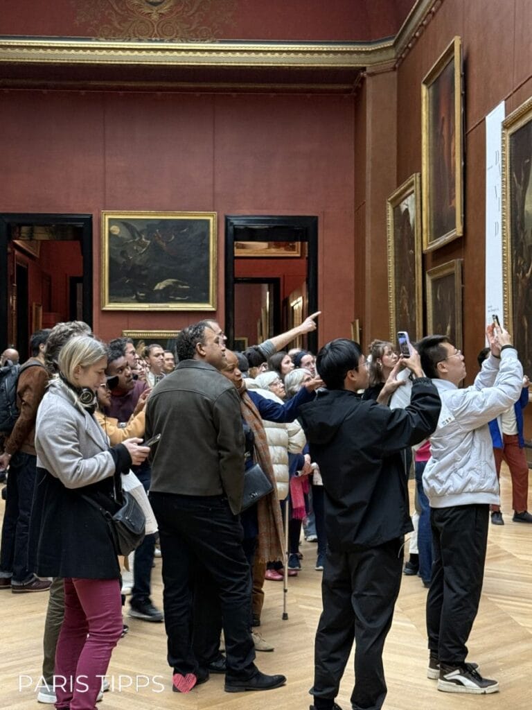 Besuchergruppe im Louvre betrachtet Gemälde an roten Wänden, einige zeigen mit dem Finger oder fotografieren die Kunstwerke. Unten links ist der Text "PARIS TIPPS ❤️" sichtbar.
