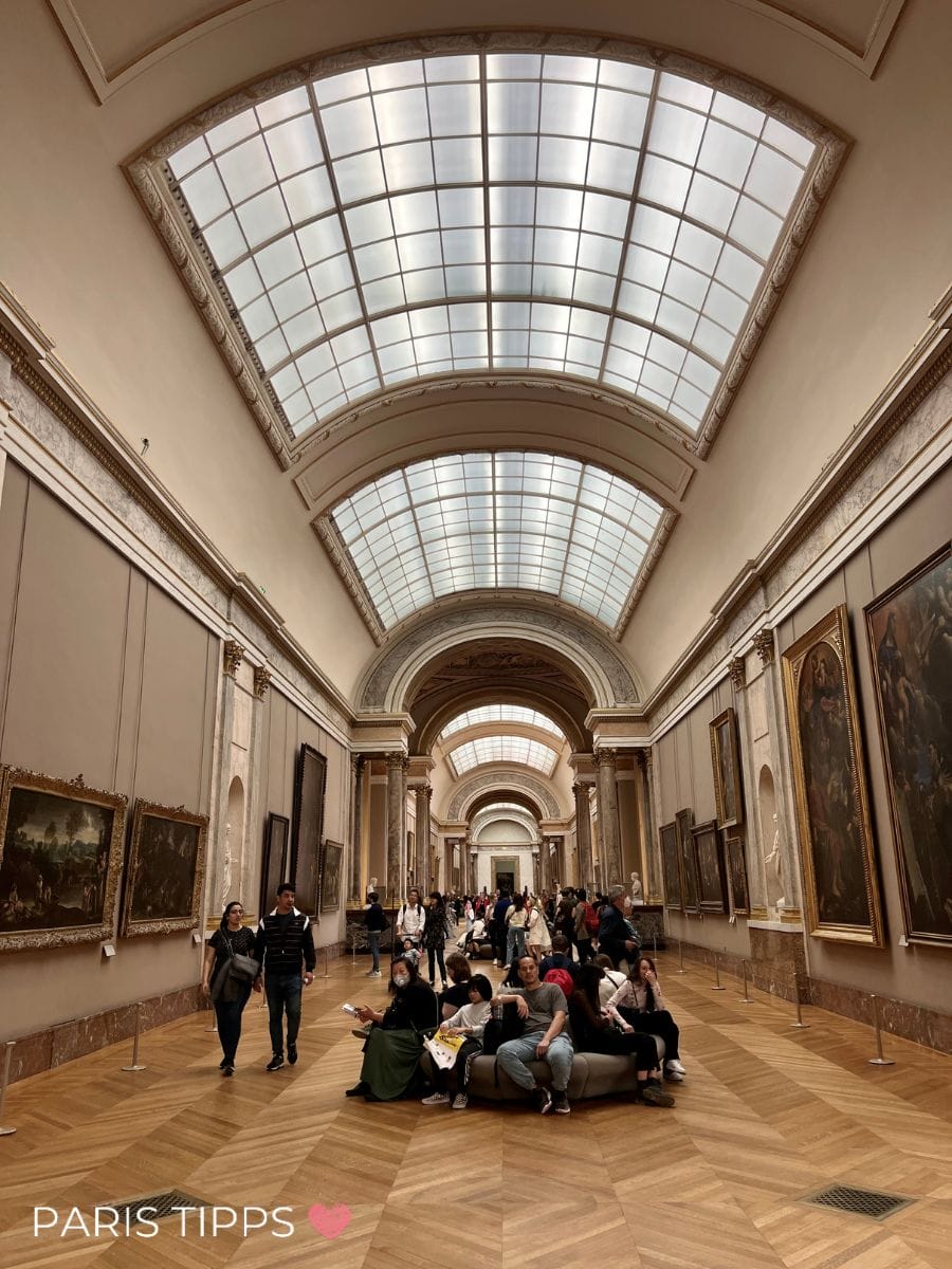 Große Galerie im Louvre mit gewölbter Glasdecke und langen Reihen klassischer Gemälde an den Wänden. Zahlreiche Besucher sitzen und gehen durch den Saal, der Teil des Rundgangs nach dem eintritt louvre ist, und unten links steht "PARIS TIPPS ❤️".