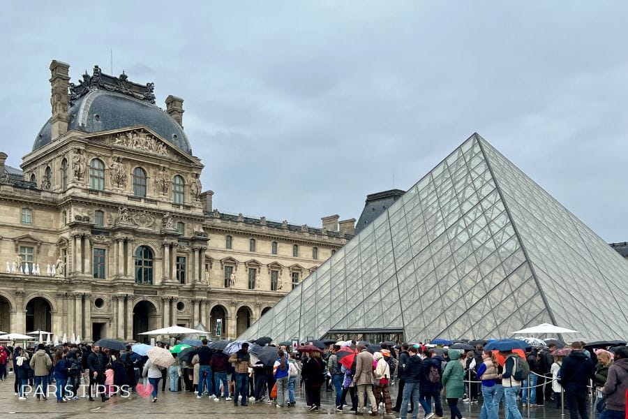 Lange Warteschlange von Besuchern mit Regenschirmen vor der Glaspyramide des Louvre an einem grauen Regentag. Die Szene zeigt den Andrang am Eingang und die typische Situation rund um den eintritt louvre, mit dem Text "PARIS TIPPS ❤️" unten links.