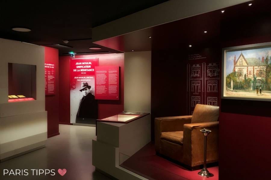 Ein Ausstellungsraum im Musée de la Libération de Paris, gewidmet Jean Moulin, mit roten Wänden, Informationstafeln und beleuchteten Vitrinen. Rechts steht ein brauner Sessel neben einem kleinen Tisch, und ein Gemälde eines Gebäudes hängt an der Wand. 