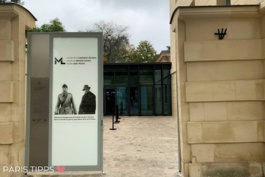 Eingang zum Musée de la Libération de Paris – Musée du Général Leclerc – Musée Jean Moulin, mit einem Schild, das zwei historische Figuren zeigt. Der moderne Glas-Eingangsbereich steht in Kontrast zur historischen Sandsteinarchitektur der Umgebung. Im Vordergrund ist eine gepflasterte Fläche zu sehen, und oben links steht 'Paris Tipps' mit einem rosa Herz.