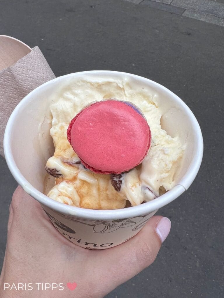 Becher mit cremigem Eis, übergossen mit Espresso (Affogato), garniert mit einem pinken Macaron, gehalten von einer Hand auf einer Pariser Straße