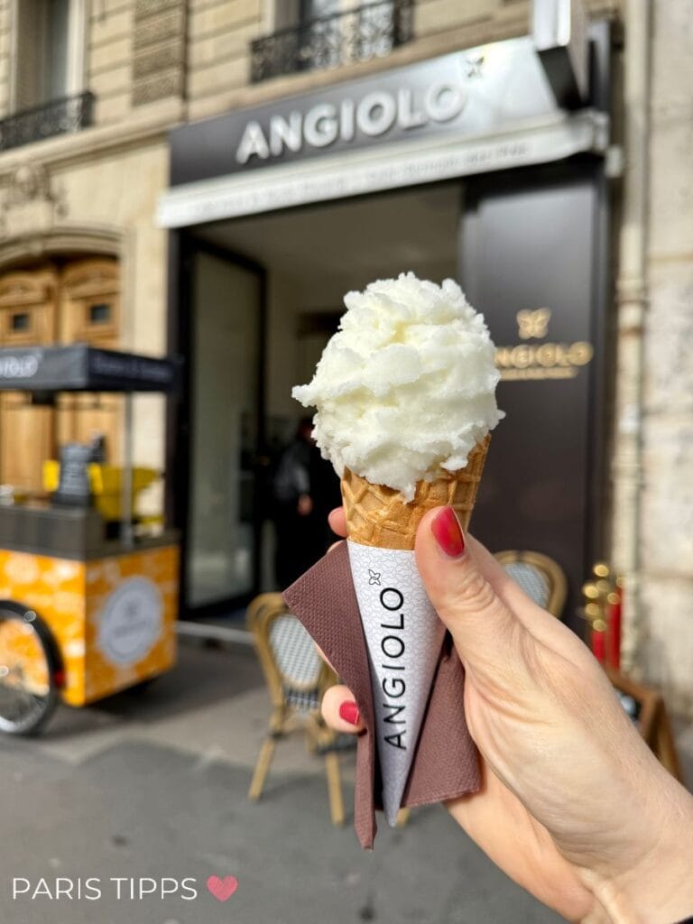 Hand hält eine Eiswaffel mit einer hellen Sorbet-Kugel vor der Eisdiele Angiolo Gelato im 6. Arrondissement in Paris. Im Hintergrund ist die Ladenfassade und ein gelber Eiswagen zu sehen.