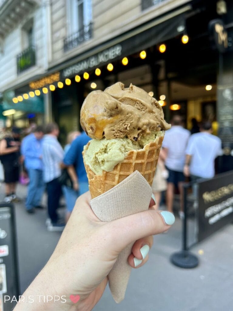 Hand hält eine Waffel mit zwei Kugeln Eis in Pistaziengrün und Karamellbraun vor der Eisdiele Glaces Moustache in Paris, im Hintergrund Menschen in der Warteschlange.