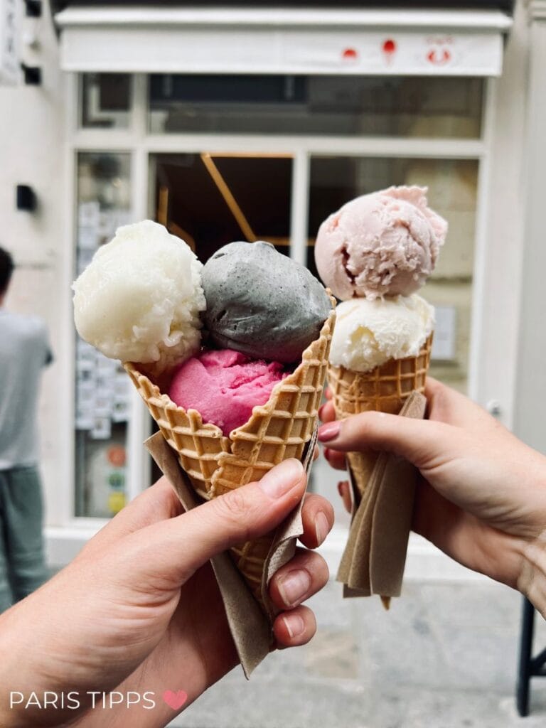Zwei Waffelhörnchen mit je drei Kugeln Eis in verschiedenen Farben – darunter weiß, pink, grau und rosa – werden vor einer Pariser Eisdiele in die Kamera gehalten.