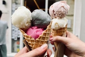 Zwei Hände halten Eiswaffeln mit jeweils drei Kugeln in verschiedenen Farben – darunter Sorbet in Weiß, Rosa, Pink und eine auffällige graue Sorte, vermutlich schwarzer Sesam, vor einer Pariser Eisdiele.
