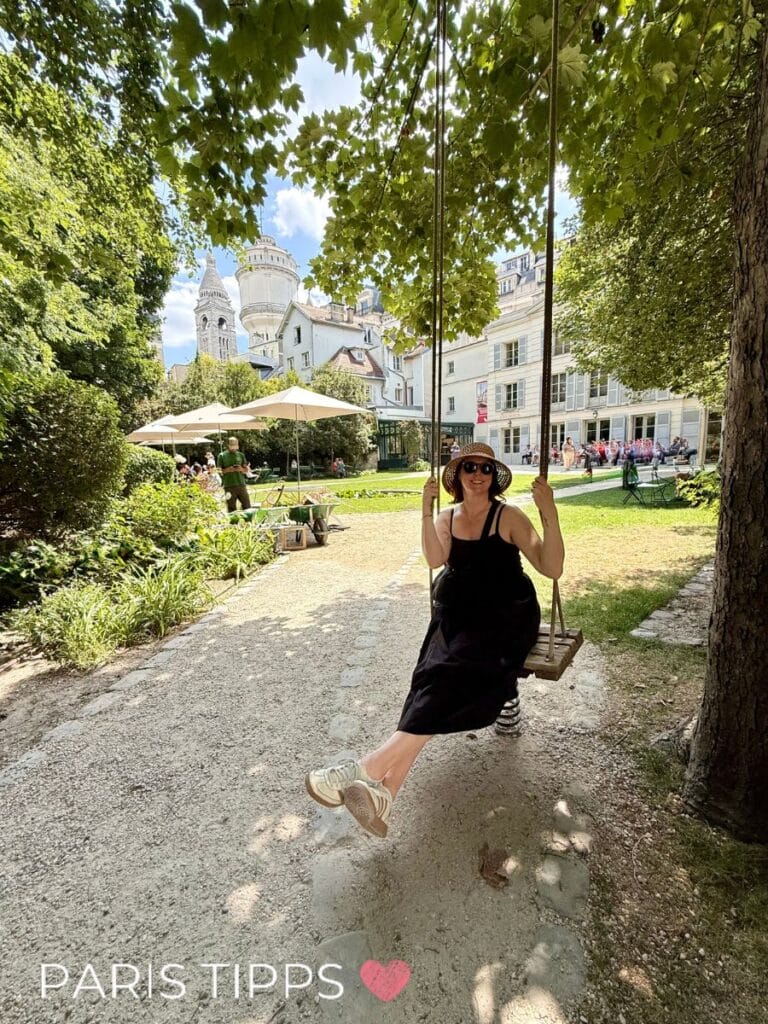 Eine Frau mit Sonnenhut und Sonnenbrille sitzt auf einer Holzschaukel unter schattigen Bäumen. Im Hintergrund ist der Garten des Musée de Montmartre zu sehen, mit Sonnenschirmen, Besuchern und der weißen Kuppel der Sacré-Cœur-Basilika. Die Stimmung ist sommerlich und entspannt. Schriftzug „PARIS TIPPS“ mit Herzsymbol am unteren Bildrand.