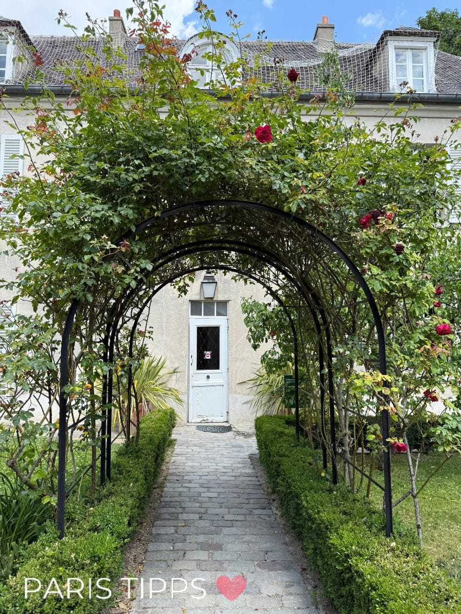 Ein bepflanzter Rosenbogen mit roten Blüten führt über einen gepflasterten Weg zu einer weißen Tür. Links und rechts säumen Buchsbaumhecken den Weg. Das Gebäude im Hintergrund hat ein helles Mauerwerk und ein Dach mit Gaubenfenstern. Unten im Bild ist der Schriftzug „PARIS TIPPS“ mit einem Herzsymbol.