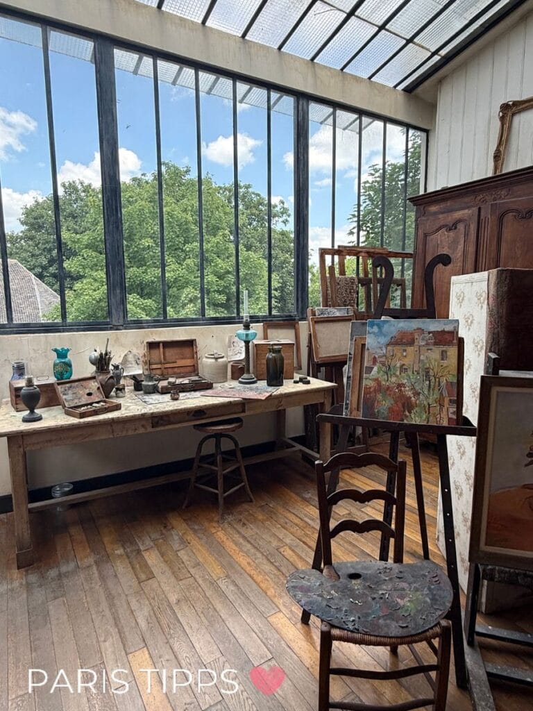 Ein historisches Künstleratelier mit großen Fenstern unter dem Dach. Holzfußboden, Malutensilien, Staffeleien und Gemälde sind im Raum verteilt. Auf dem Arbeitstisch stehen alte Pinsel, Flaschen und Farbtöpfe. Die Szene wirkt authentisch und lebendig. Schriftzug „PARIS TIPPS“ mit Herzsymbol unten im Bild.
