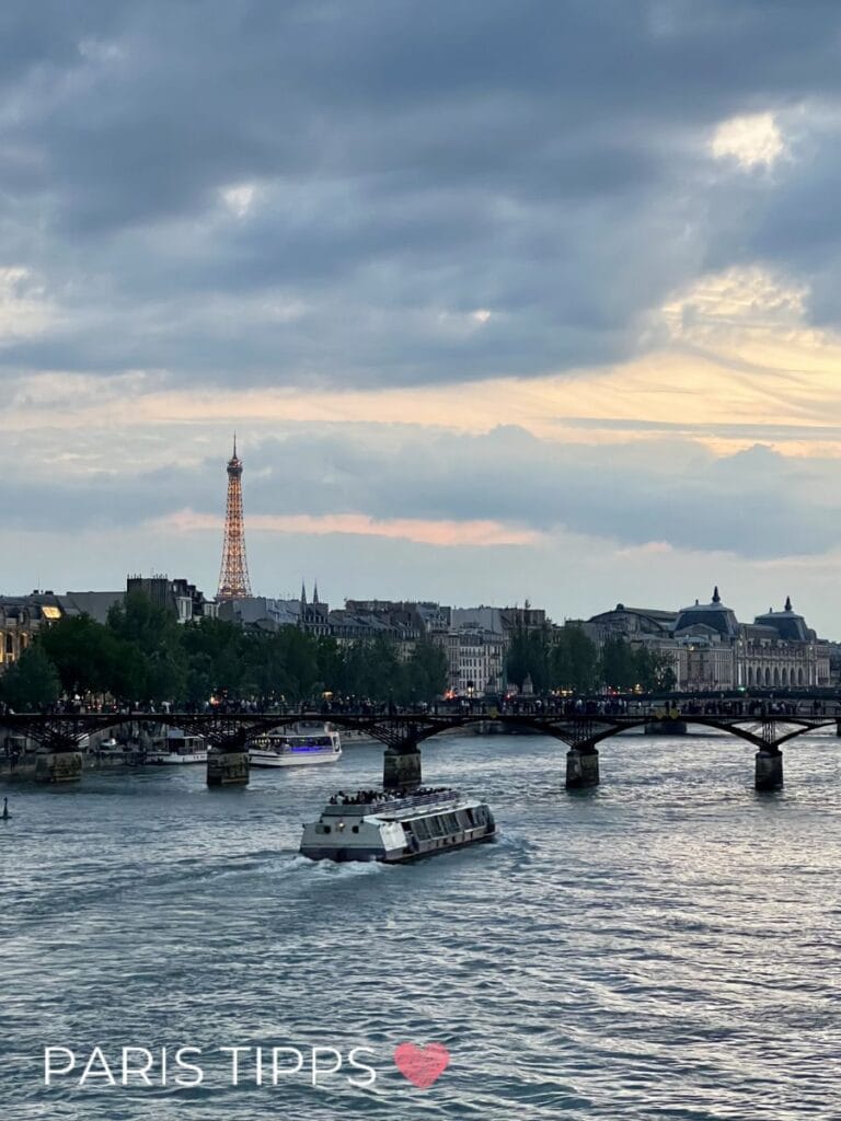 Ein Ausflugsboot fährt bei Sonnenuntergang über die Seine mit Blick auf den beleuchteten Eiffelturm und eine Brücke voller Menschen. Der Himmel ist wolkig mit warmem Licht am Horizont und spiegelt sich im Wasser. Unten links steht "PARIS TIPPS " und vermittelt die Stimmung einer abendlichen Bootsfahrt in Paris.