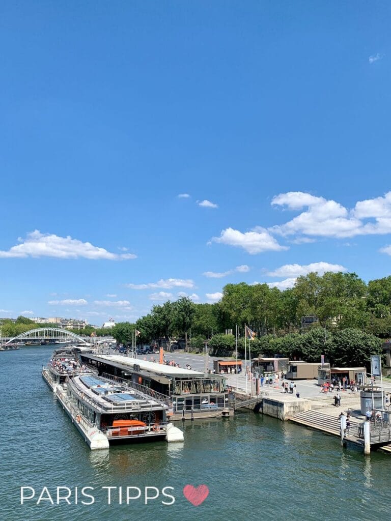 Mehrere Ausflugsboote liegen an einem Anleger an der Seine während Menschen ein- und aussteigen. Im Hintergrund sind Bäume, Gebäude und eine Brücke unter blauem Himmel zu sehen. Der Text "PARIS TIPPS ❤️" unten links verweist auf Tipps für eine Bootsfahrt auf der Seine.