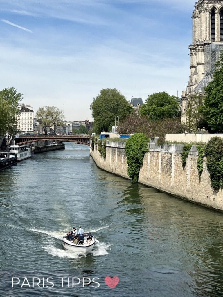 Ein kleines Motorboot fährt über die Seine entlang einer steinernen Ufermauer mit Blick auf historische Gebäude und Notre Dame. Im Hintergrund spannt sich eine Brücke über den Fluss während das Wasser leichte Wellen zieht. Unten links steht "PARIS TIPPS ❤️" und zeigt eine ruhigere Perspektive der Bootsfahrt in Paris.