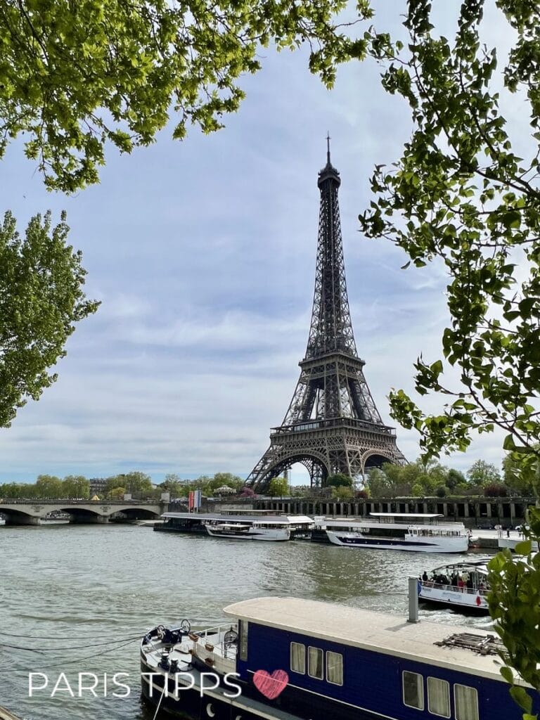 Der Eiffelturm ragt über die Seine während Boote darunter vorbeifahren und von grünen Bäumen eingerahmt werden. Eine Brücke verbindet die Ufer und ergänzt die klassische Paris Szenerie. Unten links steht "PARIS TIPPS" und zeigt ein Highlight einer Bootsfahrt auf der Seine.