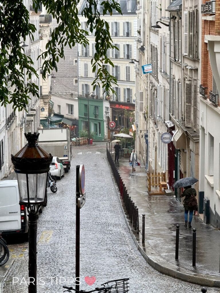Schmale Pariser Straße im Regen mit nassem Kopfsteinpflaster und Menschen mit Regenschirmen. "PARIS TIPPS ♥" steht unten im Bild und zeigt eine typische Szene von Paris im Regen.