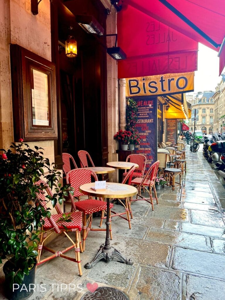 Außenbereich eines Bistros in Paris im Regen mit roten Markisen und leeren Tischen auf nassem Gehweg. Das Schild zeigt "Bistro" und "Les Jalles" und darunter eine Speisekarte.