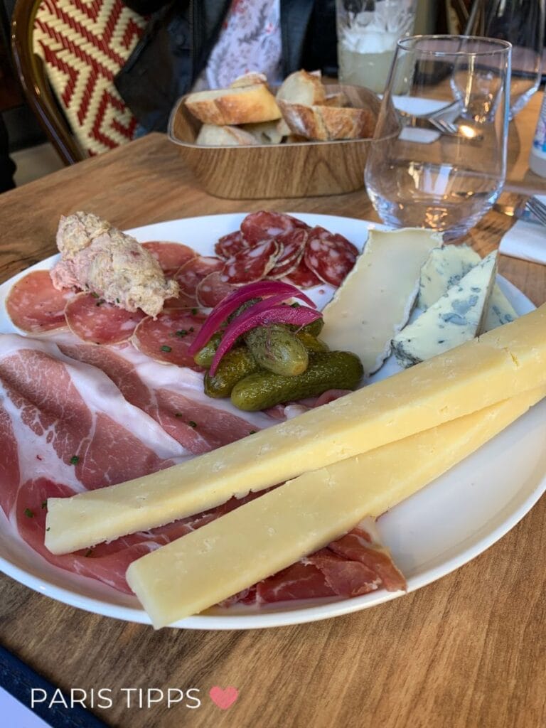 Teller mit Käse, Schinken, Salami, Essiggurken und Brot auf einem Holztisch in einem Pariser Café. "PARIS TIPPS ♥" steht unten im Bild und zeigt Essen in Paris im Regen als gemütliche Pause.