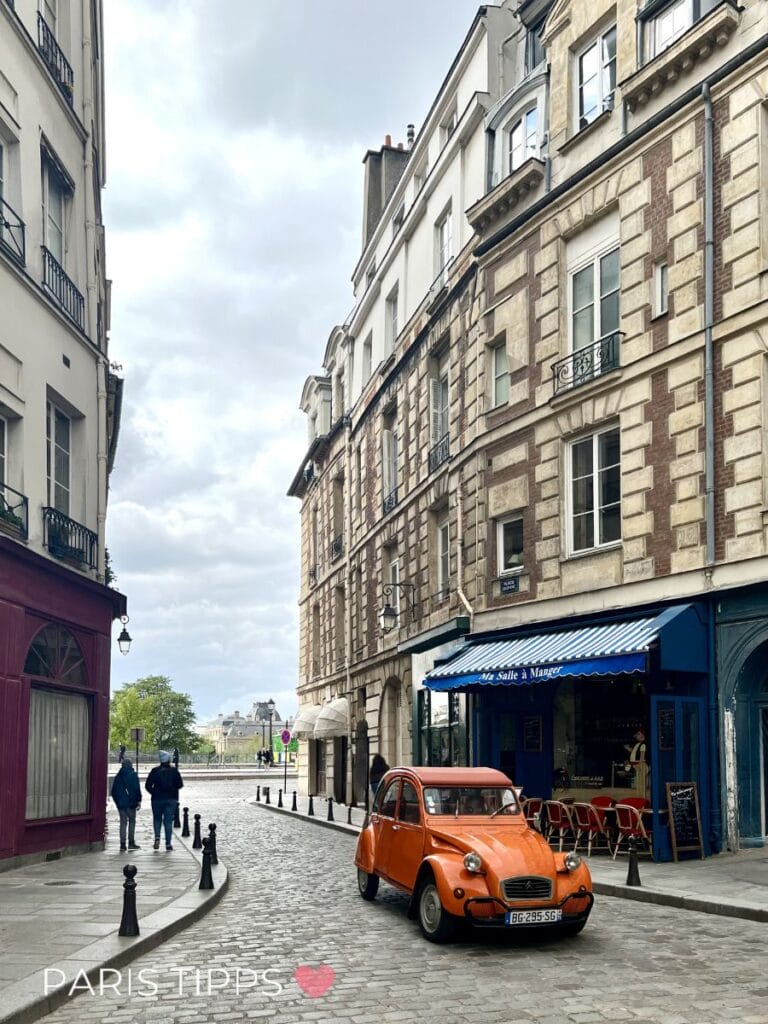 Orangefarbenes Auto fährt durch eine Pariser Straße im Regen mit alten Gebäuden und einem Café im Hintergrund. "PARIS TIPPS ♥" steht unten im Bild und zeigt eine ikonische Szene von Paris im Regen.