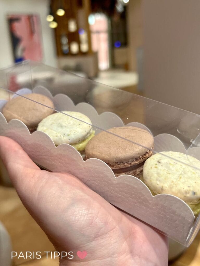 Hand hält eine Schachtel mit bunten Macarons in einem Café in Paris im Regen. "PARIS TIPPS ♥" steht unten im Bild und zeigt süße Leckereien bei Regen in Paris.